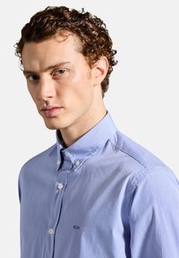 Camicia azzurra a righe con bottoni e colletto, con bottoni bianchi e un piccolo logo sul lato sinistro. Tessuto dalla texture liscia.