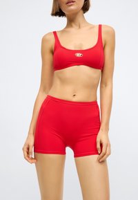 Κόκκινο σετ μπικίνι δύο τεμαχίων με κοντό τοπ και ψηλόμεσα σορτς, κατασκευασμένο από λείο ύφασμα με απαλό γυαλιστερό φινίρισμα και λεπτομέρεια λογότυπου.