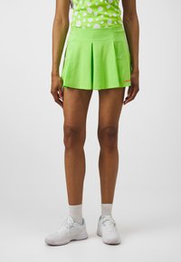 Jupe de tennis plissée verte en tissu texturé avec poches. Associée à un haut à pois colorés et des chaussures de sport blanches.