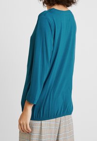 Teal langærmet top med rund halsudskæring og elastisk talje. Glat stof med en afslappet pasform, parret med en mønstret nederdel.