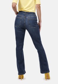 Jeans acampanados de mezclilla azul oscuro, con una cintura alta, dos bolsillos traseros y un dobladillo limpio. Combinados con una blusa amarilla y tacones amarillos.