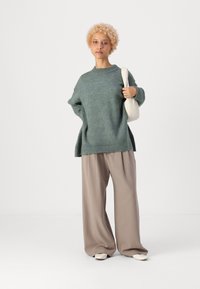 Maglione in lana taglia oversize color turchese con scollo a coste, abbinato a pantaloni beige a gamba larga e scarpe bianche a punta. Una semplice borsa bianca completa il look.
