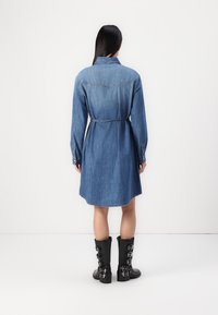 Denim srajčna obleka v srednji modri barvi z dolgimi rokavi, ovratnikom in pasom, z ukrivljenim robom in preprostimi šivi. Črne gležnarje z zaponkami.