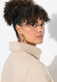 Beige Oberteil mit hohem Kragen und weichem Stoff. Eine Frau mit lockigem Haar trägt große goldene Creolen und steht vor einem weißen Hintergrund.