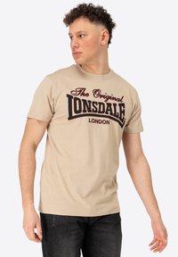 Beidzis kokvilnas t-krekls ar īsām piedurknēm, uz kura priekšpuses ir liels melns un sarkans grafiskais teksts "The Original LONSDALE LONDON".