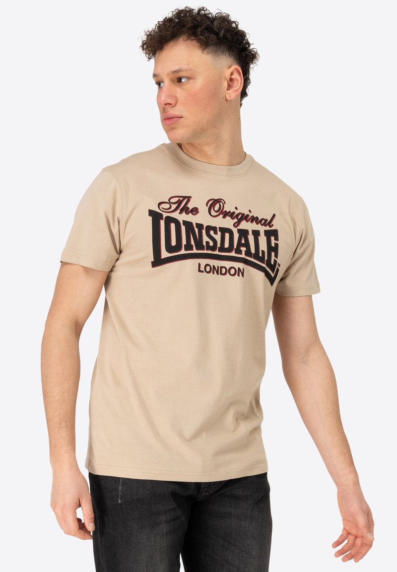 Beidzis kokvilnas t-krekls ar īsām piedurknēm, uz kura priekšpuses ir liels melns un sarkans grafiskais teksts "The Original LONSDALE LONDON".