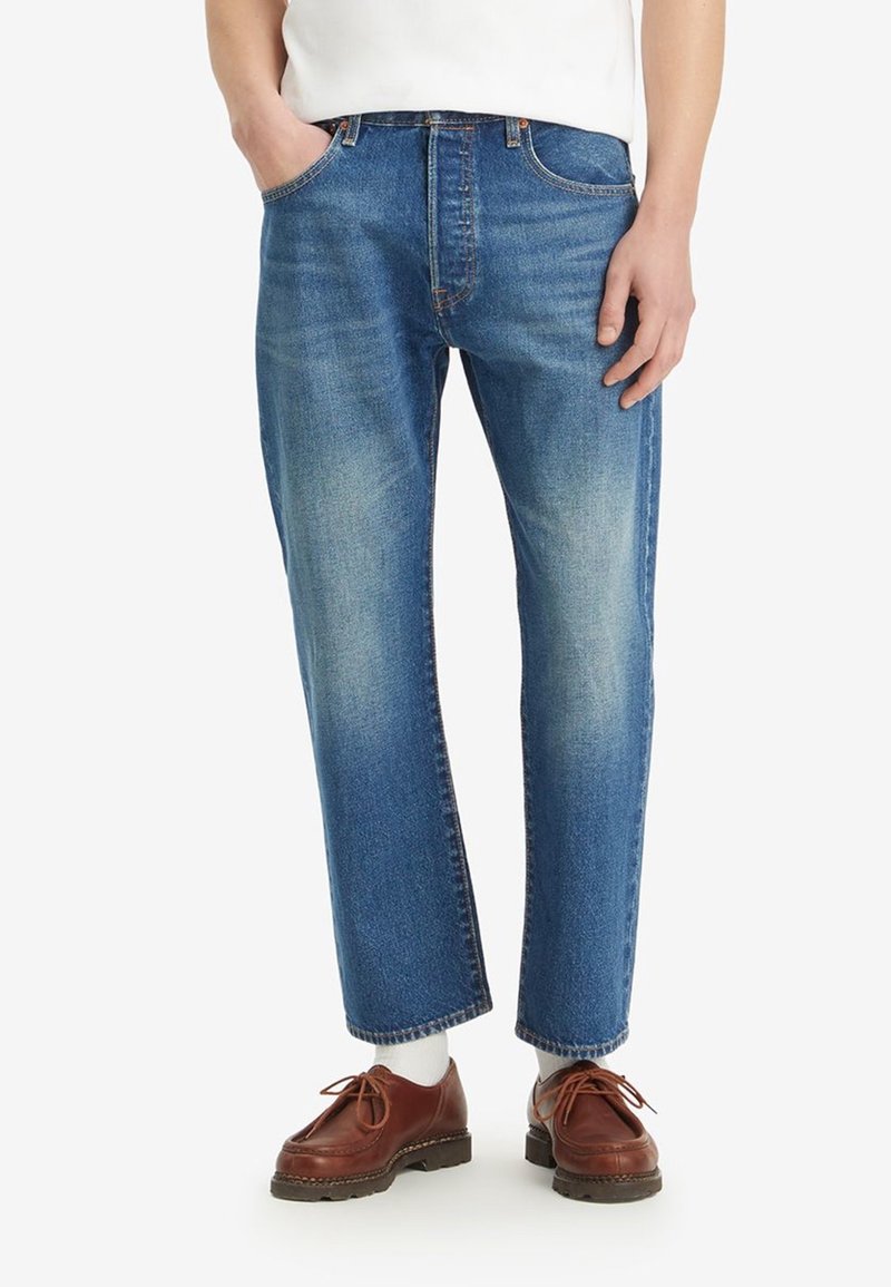 Homme portant un jean droit bleu moyen, des chaussettes blanches, des chaussures en cuir marron à lacets, et une chemise blanche rentrée avec les mains dans les poches.