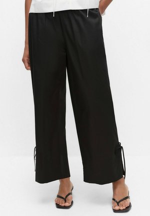 Sorte bukser med vide ben og snøre ved kanterne, båret med sorte thong-sandaler, der viser nedre torso og ben mod hvid baggrund.