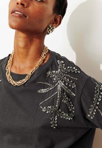 T-shirt en coton noir ornée d'une broderie florale argentée en perles et fil sur l'épaule, superposée avec plusieurs colliers en chaîne dorée.