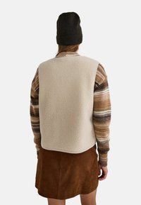 Personne portant un gilet en polaire beige, un pull à rayures marron et beige à manches longues, une jupe en daim marron et un bonnet en tricot noir, vu de dos.
