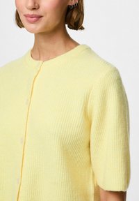 Gaiši dzeltena adīta džemperis ar apaļu kakla izgriezumu, īsām piedurknēm un pogu aizdari. Faktūra ar ribotu rakstu.