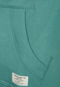 Tessuto di felpa verde acqua con tasca frontale, dettagli a coste e un'etichetta con la scritta "JACK & JONES CO.". I bordi sono rifiniti con cuciture a vista che definiscono il design.