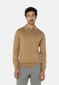 Langärmliges, hellbraunes Poloshirt aus einem weichen Stoff mit Kragen und Knopfleiste mit drei Knöpfen. Kombiniert mit grauen Hosen.