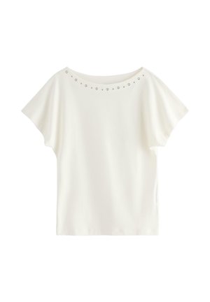 Jeune femme debout, main sur la hanche, portant un haut blanc à manches courtes, un jean bleu, des baskets beiges, tenant un petit sac à main noir.