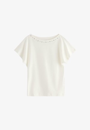 Ung kvinde står med hånden på hoften, iført hvid kortærmet top, blå jeans, beige sneakers og holder en lille sort håndtaske.