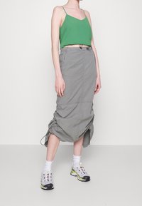 Žena v zeleném bezrukávovém topu, šedé zřasené midi sukni, bílých ponožkách a bílých teniskách s žlutými akcenty stojící na jednobarevném pozadí.