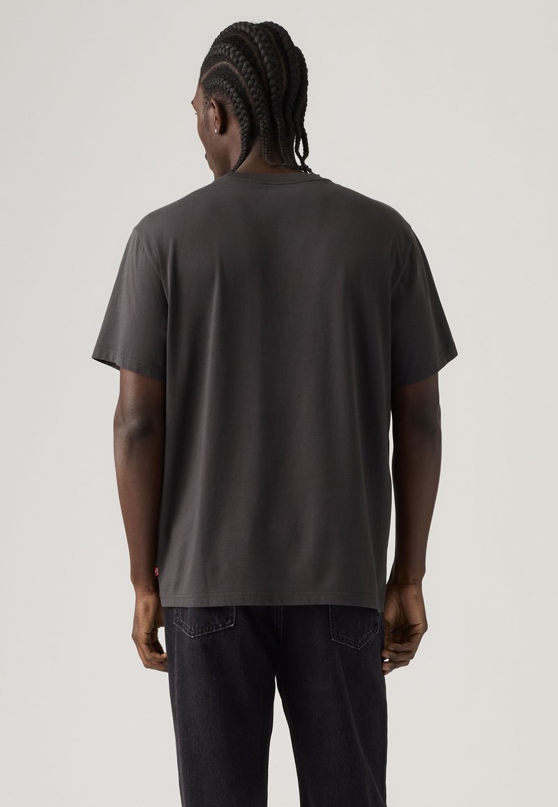 Man with braided hair wearing a plain dark gray t-shirt and dark jeans facing away against a light gray background.