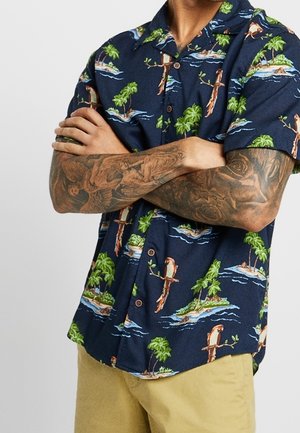 Person med krydsede arme iført en marineblå kortærmet skjorte med tropisk ø og fugleprint samt beige bukser, der viser detaljerede tatoveringer på armen.