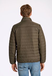 Olivgrön puffjacka med horisontellt quiltat mönster, hög krage och elastiska ärmslut. Parades med blå jeans. Minimalistisk design.