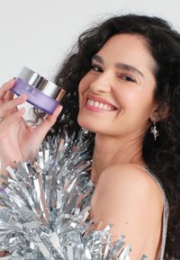 Crème violette dans un pot rond avec un couvercle argenté. Le modèle tient le pot tout en portant des franges en aluminium argenté. Texture lisse à la surface du pot.