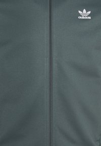 Donkergroene sportjack met een rits over de volle lengte, geribbelde textuur en een witte Adidas-logo op de linkerkant van de borst. Glad, lichtgewicht materiaal.