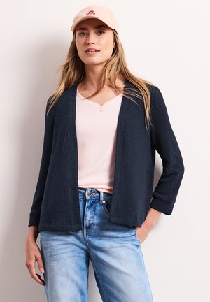 Kvinde iført en lyserød kasket, lyserød top, mørk åben cardigan og lyseblå jeans, stående op ad en ensfarvet væg.