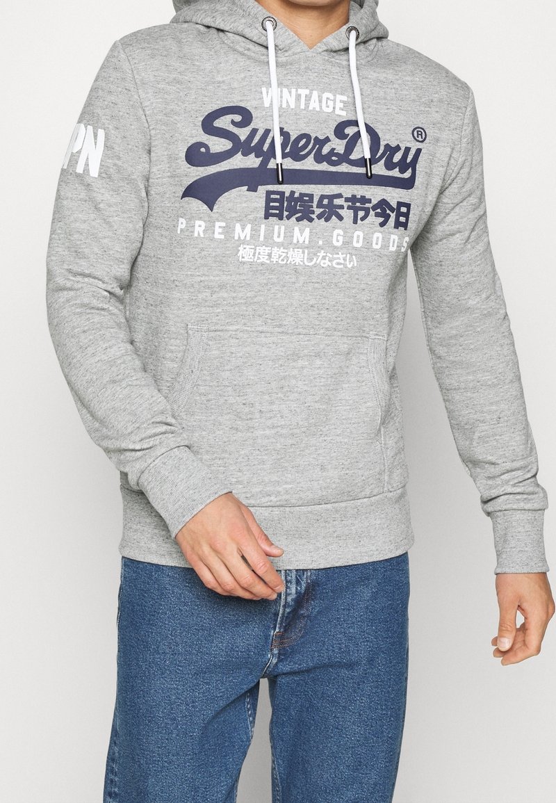 Grå hoodie tillverkad av ett mjukt material med känguruficka, vita dragsnören och marinblå tryck som har "SuperDry" och asiatiska tecken.