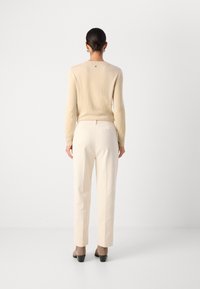 Beiger Pullover, cremfarbene Hose und taubfarbene, hochhackige Schuhe. Der Pullover hat einen gerippten Ausschnitt; die Hose hat eine strukturierte Oberfläche mit Gesäßtaschen.