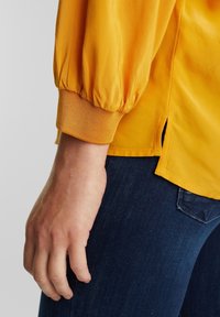 Blouse jaune à manches ballon, poignets côtelés et fentes latérales, présentée avec un jean en denim foncé. La texture semble lisse et douce.