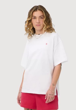Jeune personne aux cheveux ondulés portant un t-shirt blanc ample et un short rouge, debout devant un fond clair uni.