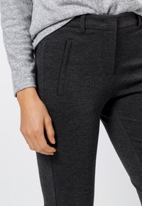 Pantalon ajusté gris foncé en tissu extensible, comportant des poches latérales et une texture lisse, associé à un pull gris clair.