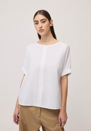 Frau trägt eine locker sitzende weiße kurzärmelige Bluse und beige Hose, mit goldenem Ear Cuff und Ringen, steht vor einem schlichten hellen Hintergrund.
