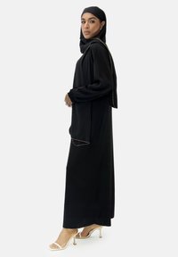 Robe noire à manches longues avec une silhouette fluide, ornée de coutures accentuées dorées et un hijab noir assorti. Le mannequin porte des talons beiges.