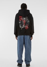 Hanorac negru cu print grafic mare pe spate, prezentând un dragon, accentuat cu text roșu. Croială oversized și manșete ribbate. Blugi bleu cu croială largă.