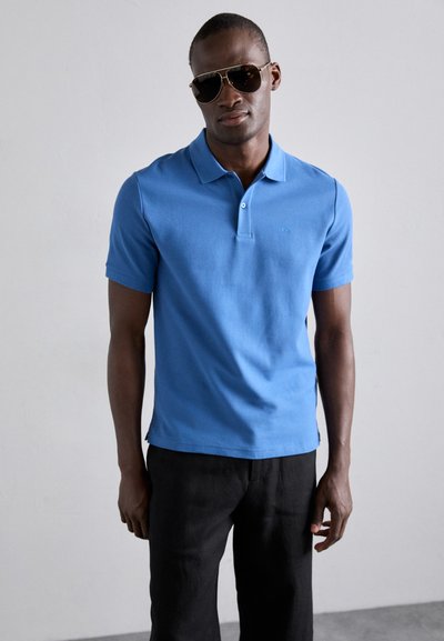 Camisa polo azul claro de algodón, con cuello, mangas cortas y un logotipo sutil. Combinada con pantalones negros y gafas de sol.