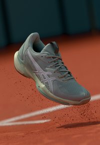 Chaussure de sport gris clair avec tige en mesh, semelle incurvée et pointe renforcée. Présente des accents roses et un logo ASICS bien visible.