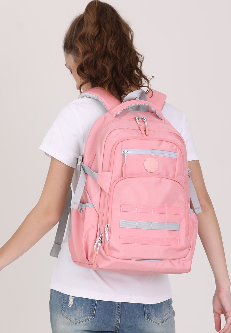 Sac à dos rose avec des accents gris, de forme rectangulaire, avec plusieurs zippers et poches latérales. Il possède des bretelles ajustables et une patch logo à l'avant.