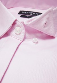 Jasnoróżowa koszula z teksturowanej tkaniny, z kołnierzykiem na guziki, dwoma białymi guzikami i metkami wewnątrz kołnierzyka.