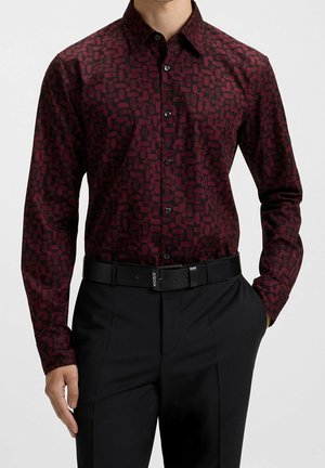 Homme portant une chemise ajustée à manches longues avec un motif abstrait rouge foncé, ceinture noire et pantalon noir, mains dans les poches.