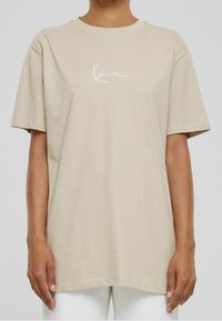 Camiseta de algodón beige con mangas cortas, escote redondo y logo en cursiva blanco "Kanie" en el pecho. Textura suave, corte relajado.