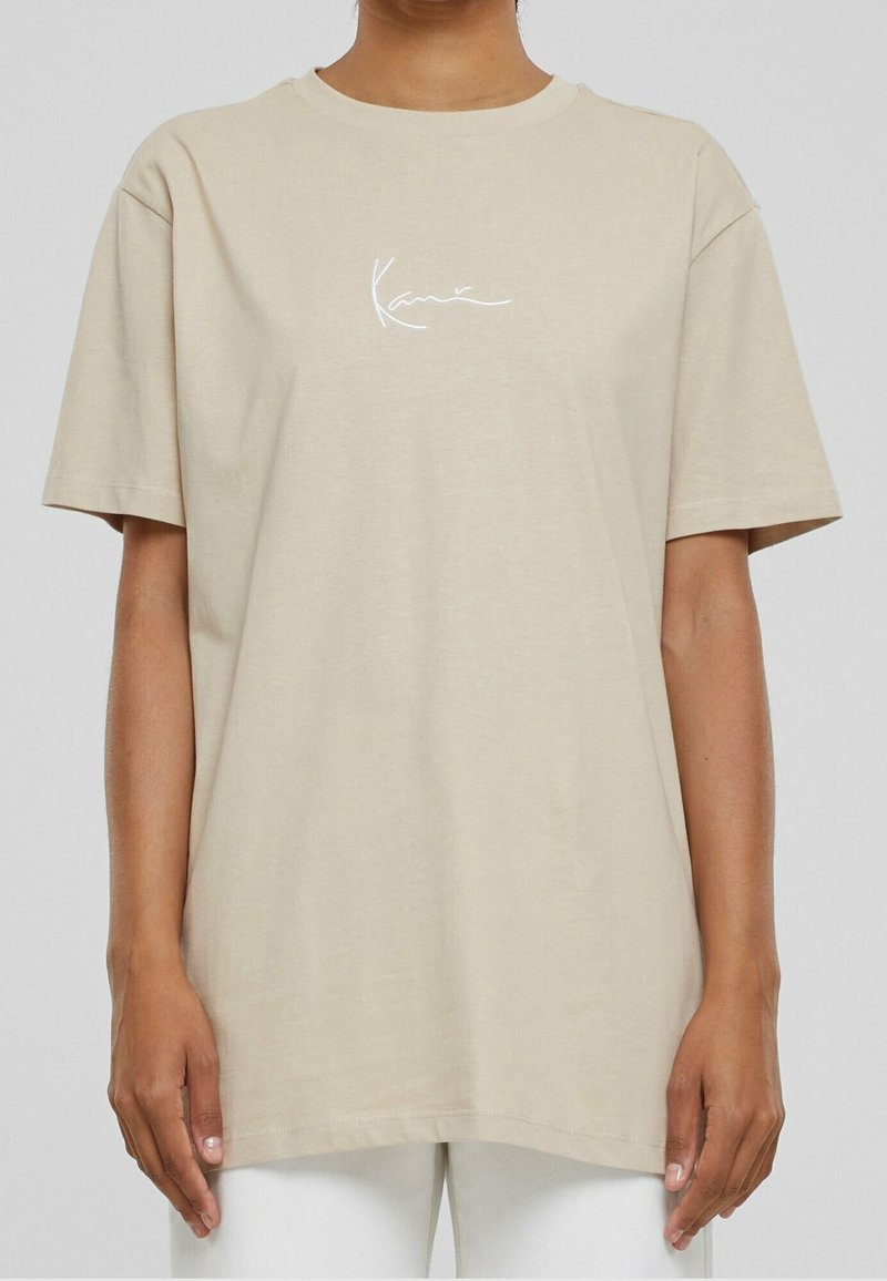 Camiseta de algodón beige con mangas cortas, escote redondo y logo en cursiva blanco "Kanie" en el pecho. Textura suave, corte relajado.