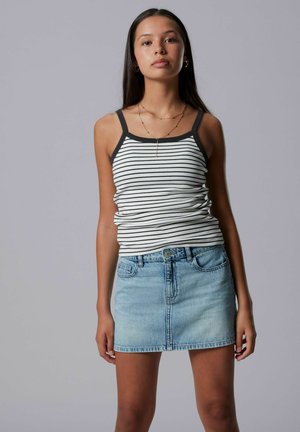 Młoda kobieta z długimi ciemnymi włosami, ubrana w czarno-biały pasiasty top na ramiączkach i jasnoniebieską dżinsową mini spódniczkę na szarym tle.