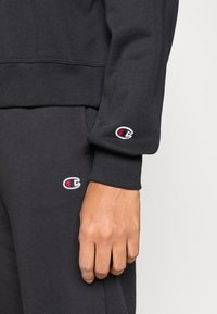 Μαύρο φούτερ με ριμπ μανσέτες, διακοσμημένο με κεντημένα λογότυπα στο μανίκι και το παντελόνι. Το ύφασμα φαίνεται μαλακό με υφή φινιρίσματος.