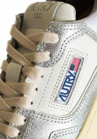 Sneaker in pelle bianca con accenti argentati, lacci beige e un'etichetta logo ricamata con colori rosso e blu. Dettagli metallici testurizzati.