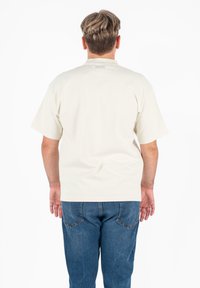 Crèmekleurig katoenen t-shirt met korte mouwen, ontspannen pasvorm, eenvoudig ontwerp en een label op de achterkant. Draagt met blauwe denim jeans.