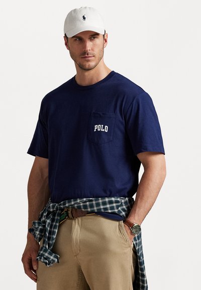 Granatowy t-shirt z białym logo "POLO" na kieszeni piersiowej. Noszony z zieloną koszulą w kratę zawiązaną w talii i beżowymi spodniami. Biała czapka.