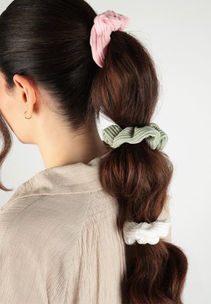 Frau mit langem, dunklem Haar, das in drei Abschnitte geteilt und mit rosa, grünen und weißen strukturierten Haargummis gebunden ist, trägt ein beiges, strukturiertes Oberteil.