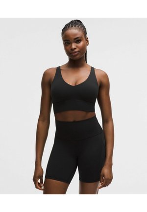 Kvinde med flettet hår iført en sort sports-bh og højtaljede sorte cykelshorts, stående mod en ensartet lys baggrund.