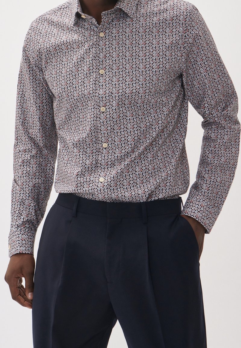 La camicia button-up presenta un piccolo e intricato motivo floreale in blu, rosso e crema su uno sfondo chiaro. Maniche lunghe e colletto standard.