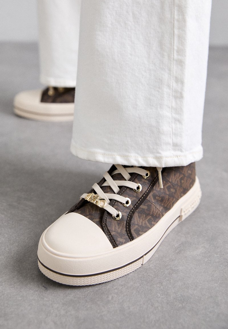 Baskets marron à motifs avec embout et lacets beiges, portées avec un pantalon blanc sur un sol gris.